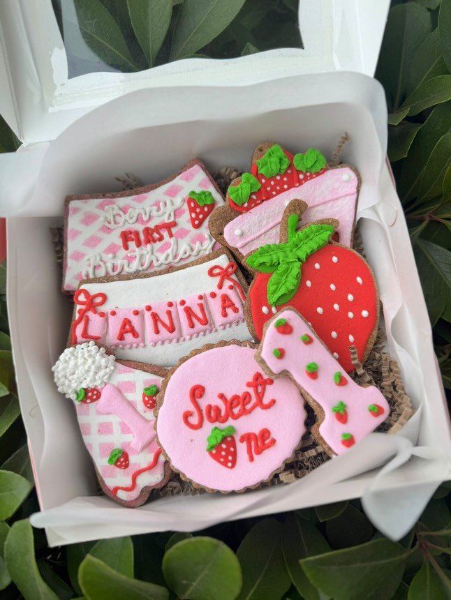 A white box filled with custom strawberry-themed cookies for a first birthday, featuring pink, red, and green icing designs including the name “LANNA,” the number “1,” and celebratory shapes. The box is styled with shredded paper and surrounded by greenery, evoking a fresh San Diego vibe.