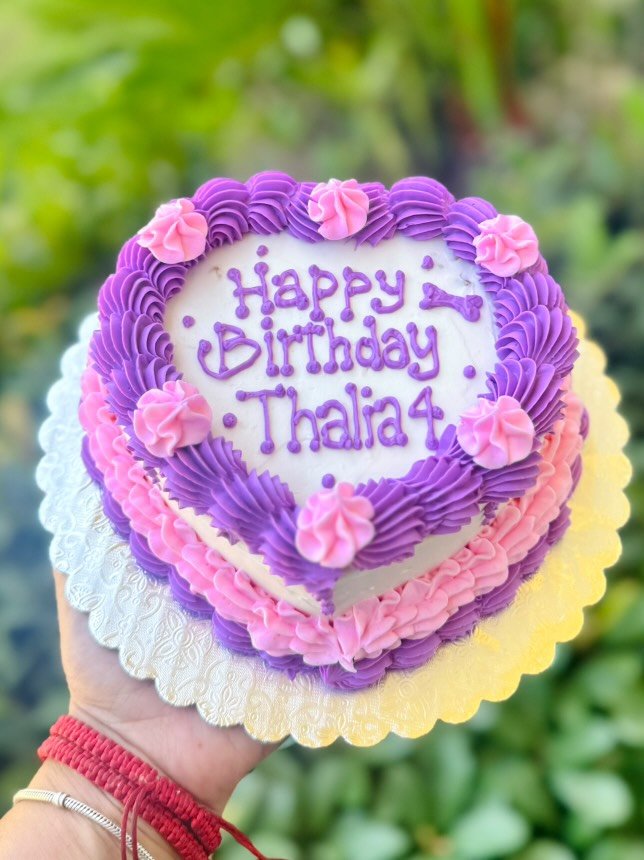 A heart-shaped birthday cake decorated with pink and purple frosting, featuring rosettes and piped borders. The cake reads “Happy Birthday Thalia 4” and is held outdoors by a hand wearing red and silver bracelets, with greenery in the background.